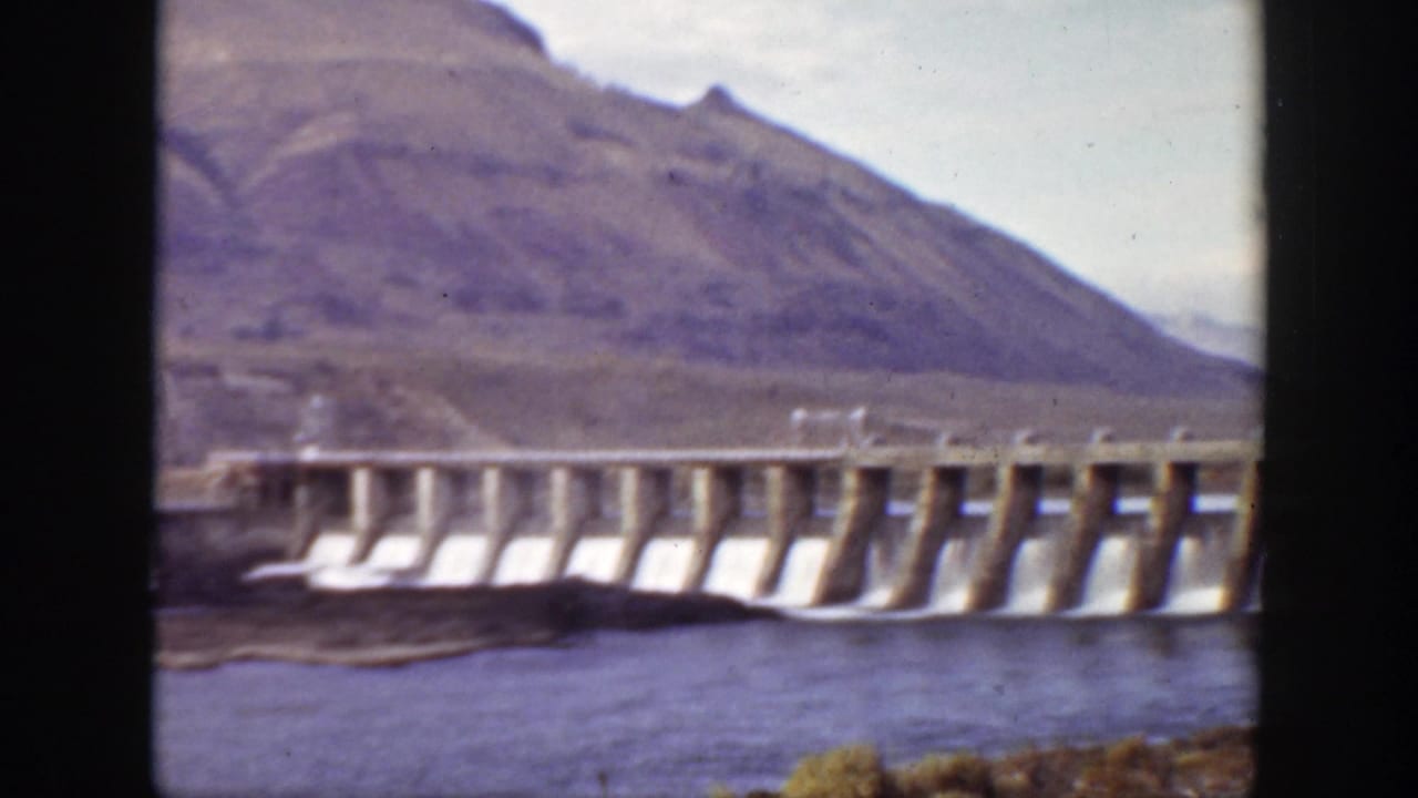 Concrete dam releasing water into a river below dry mountain slopes.