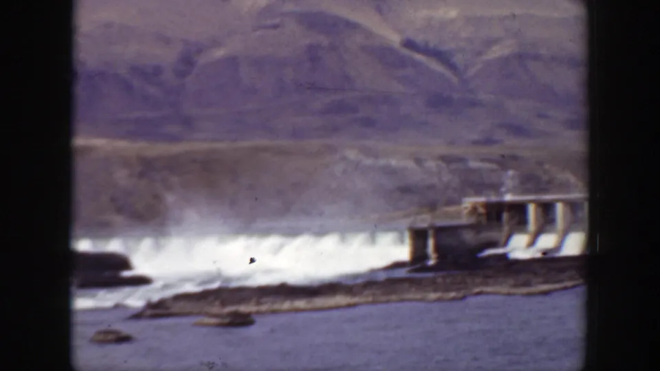 1938 California 8mm frame with the dam partly at right, white spillway water, river foreground, and arid hillside beyond.