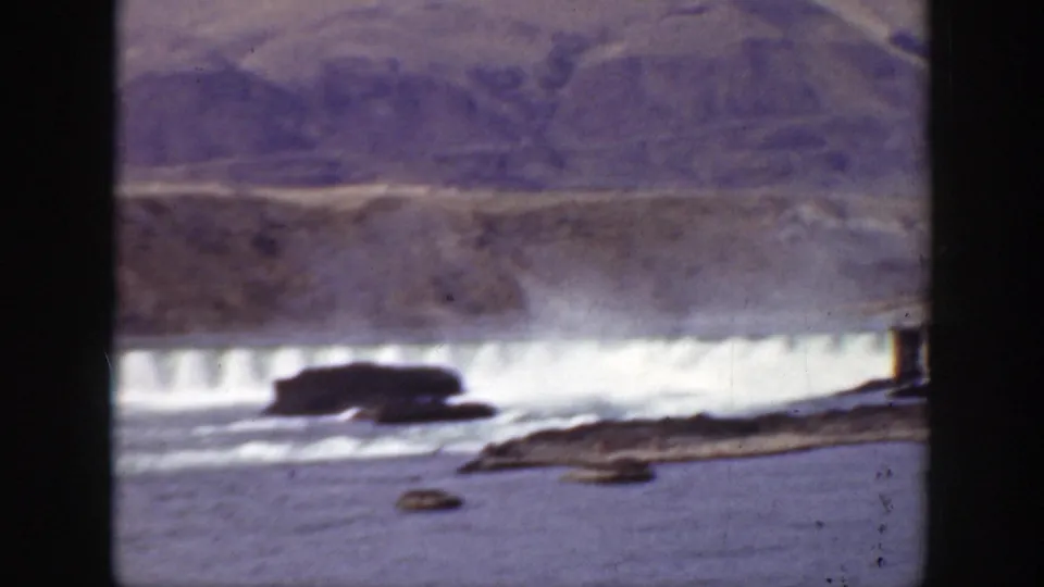 8mm home-movie frame from California showing dam spillway discharge, river rocks, and dry mountain terrain in soft archival color.