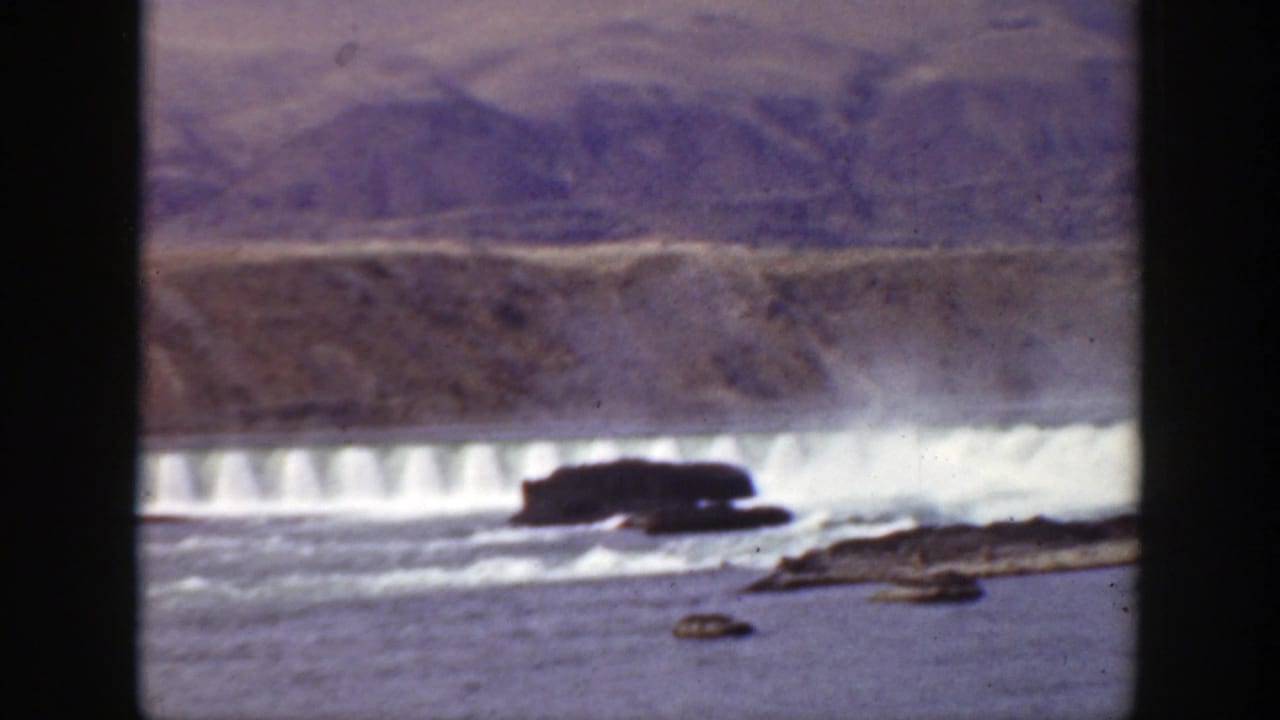 1938 California 8mm frame showing a broad river, white water below a low dam, dark rocks, and barren hills beyond.