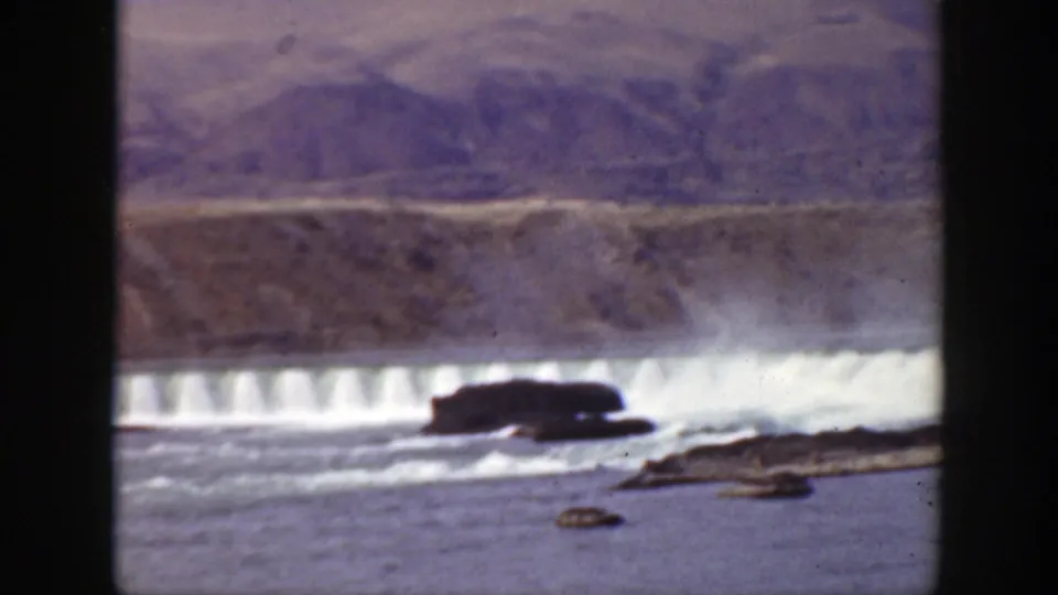1938 California 8mm frame showing a broad river, white water below a low dam, dark rocks, and barren hills beyond.