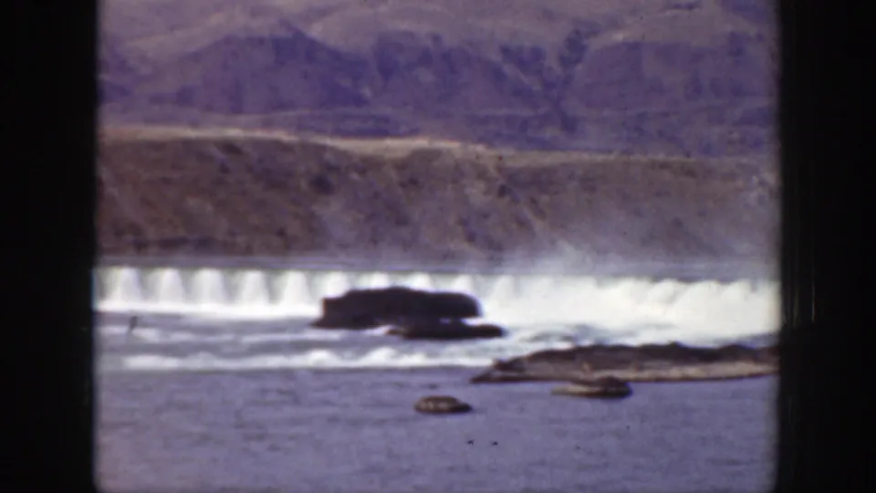 Wide 1938 California river scene with white water, a low dam or weir, dark rock outcrops, and scrub-covered hills.