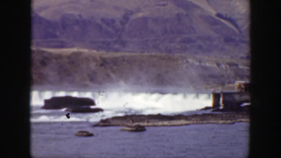 1938 California home-movie frame showing a river below a low dam, with foaming water, rocks, and a small island in front.