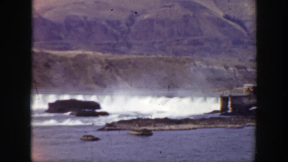 Final 1938 California 8mm frame of white water below a low river dam, with dark rocks, a small island, and dry hills behind.