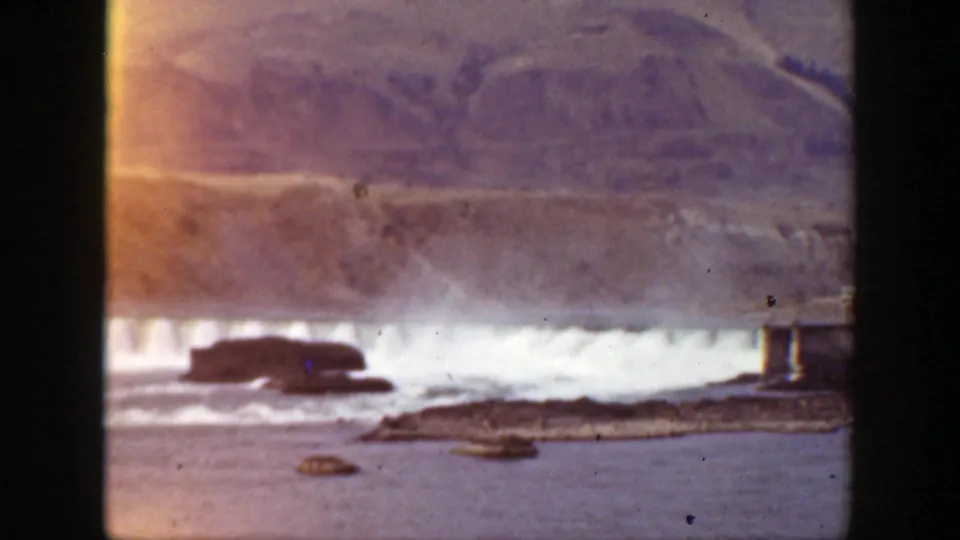 Faded 1938 California 8mm view of turbulent water below a dam, with hills and rocky outcrops visible beyond the spillway.