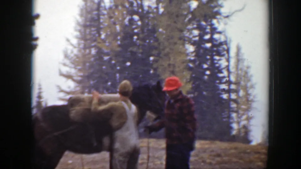 A man in overalls secures a bulky load on a dark horse as another man assists in the forested 1938 California scene.