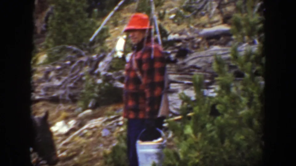 1938 California frame of a man in a red hat and plaid shirt standing near a bucket in mountain brush.