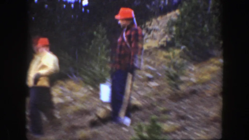A man in a red hat and plaid shirt stands with a shovel on a wooded California hillside as another man in a light jacket passes.