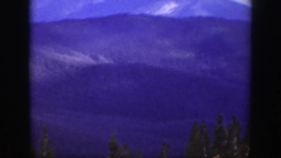 1938 California home-movie frame with distant mountain ridges, blue-purple sky, and dark evergreen treetops along the lower edge.