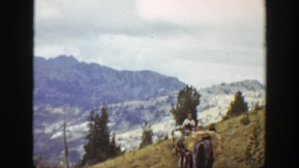 Pack animals and a rider appear near the lower hillside, with conifers and hazy mountain ridges in the background.
