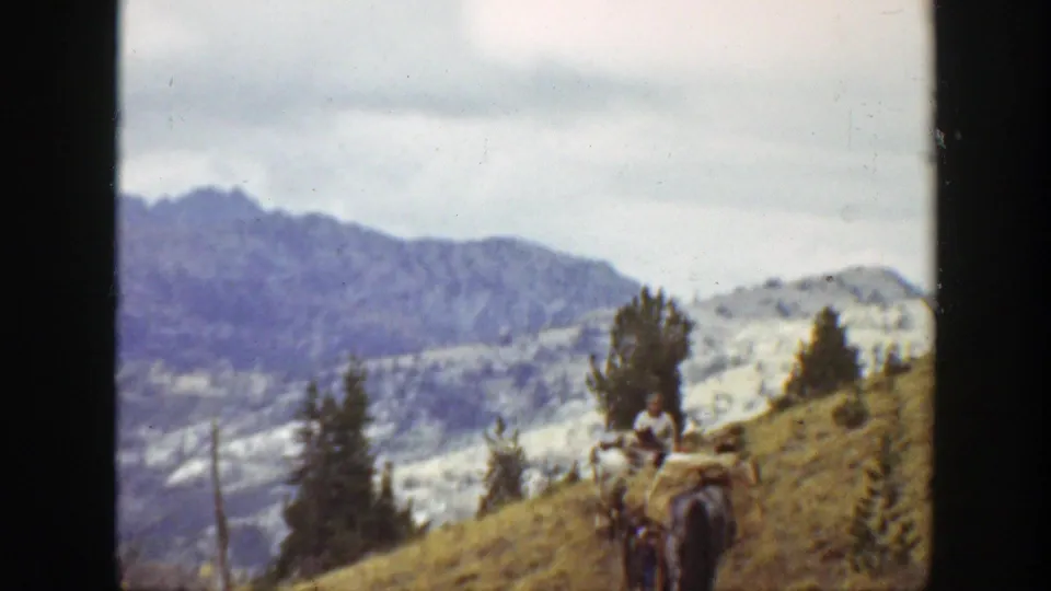 1938 California home-movie frame showing a rider on a pack animal on a mountain slope with conifers and snow-dusted peaks beyond.