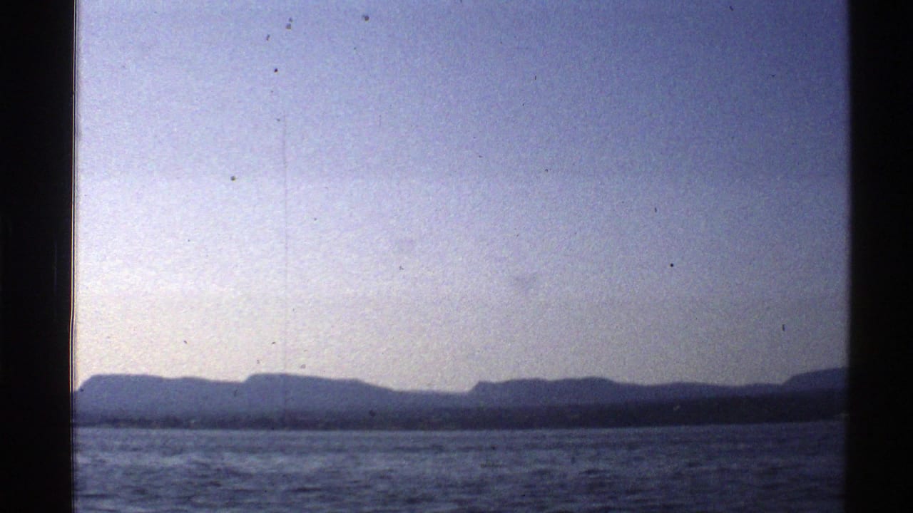 1976 Norway 8mm frame showing a lake surface and distant mountain silhouette beneath a pale blue sky.