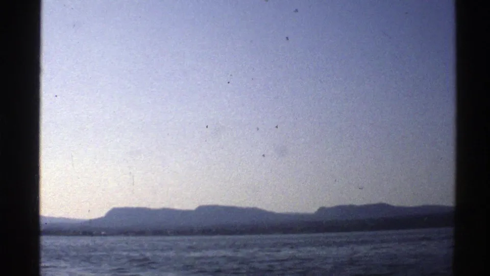 1976 Norway home-movie frame looking across rippled water toward a low, shadowed mountain range.