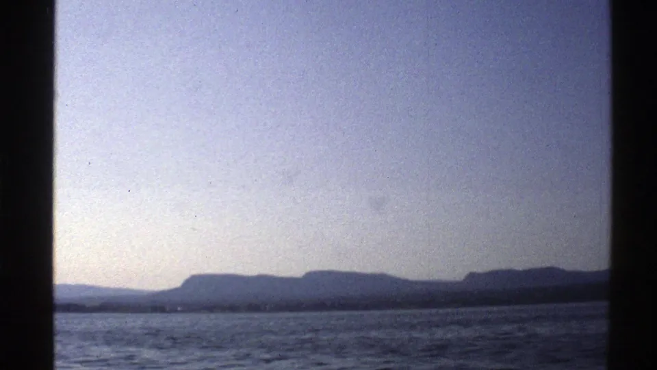 1976 Norway 8mm frame showing a broad mountain silhouette beyond lake water and a pale, lightly hazed sky.