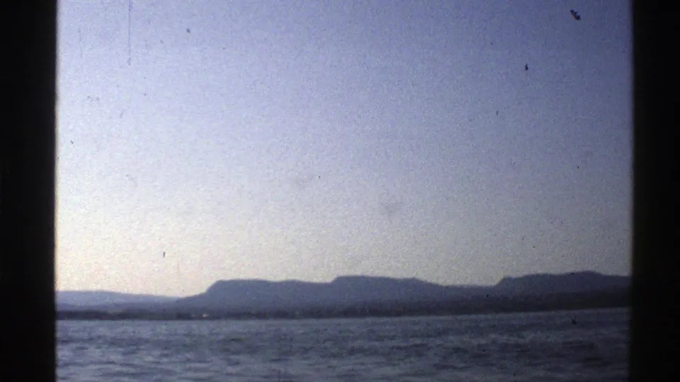 1976 Norway 8mm frame showing rippled open water and a distant mountainous shoreline beneath soft pale light.