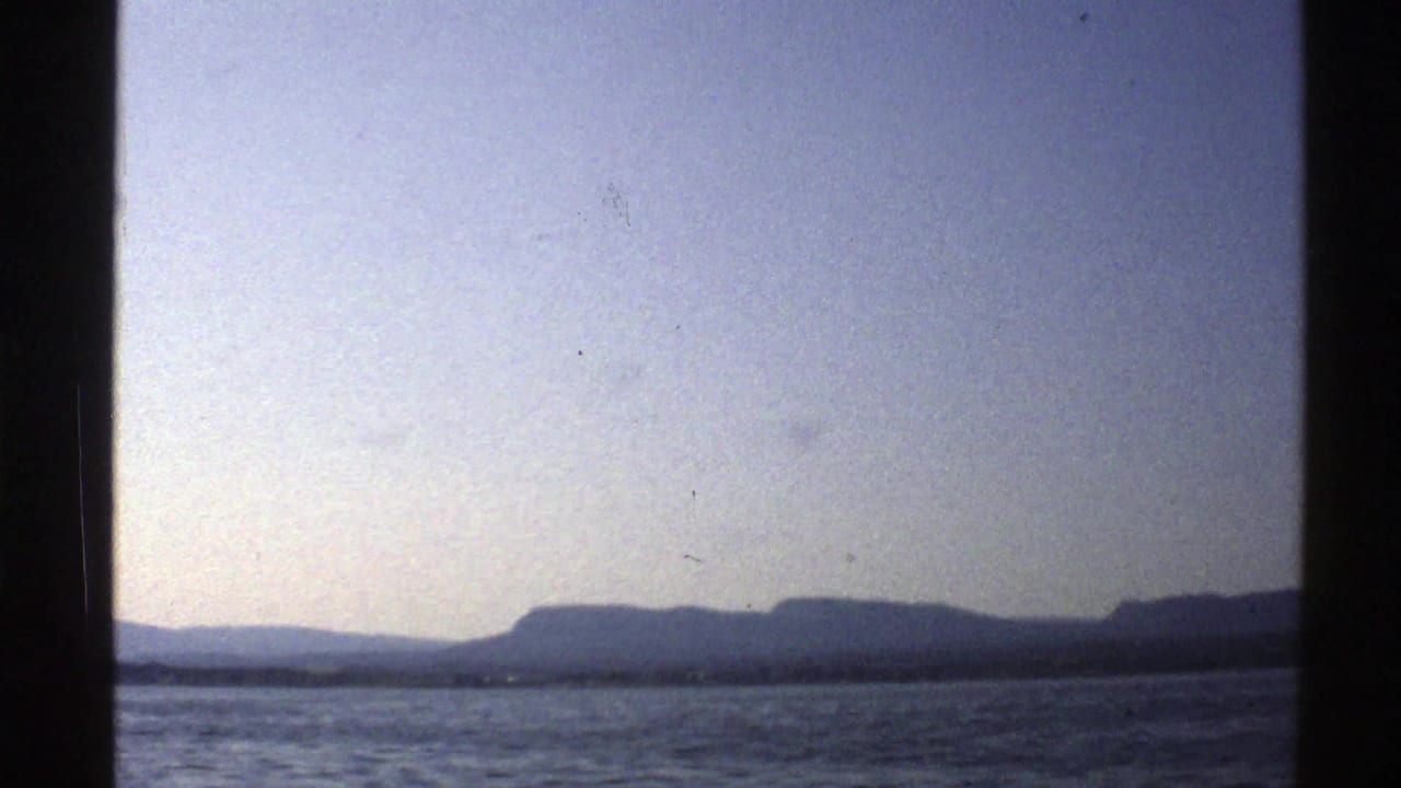 Rippling water stretches toward a dark mountainous horizon beneath a pale blue sky.