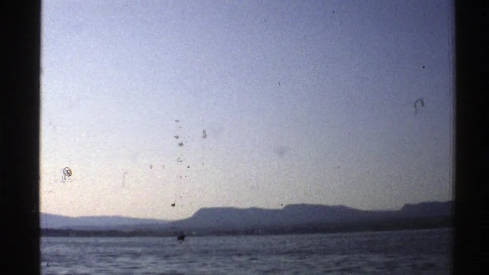 1976 Norway 8mm frame of rippled water and a far mountainous shoreline under soft, lightly marked sky.
