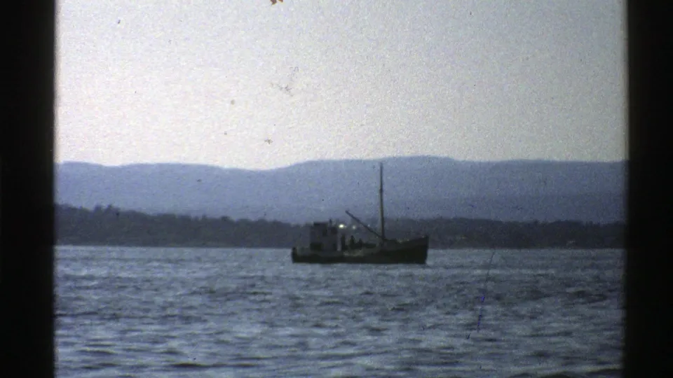 Norway coastal home-movie frame with a small boat on open water, distant shore, and muted blue-gray mountains.