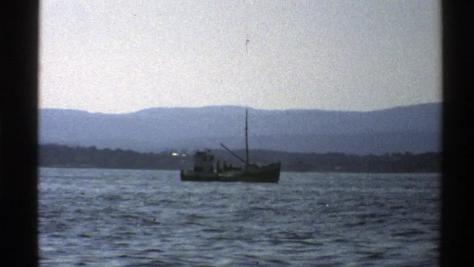 1976 Norway 8mm frame showing a small fishing boat crossing open water before a hazy mountain shoreline.