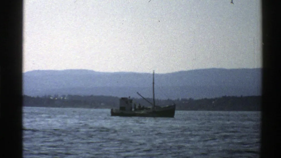Vintage 8mm view of a small Norwegian fishing boat moving across calm water before a soft mountain horizon.
