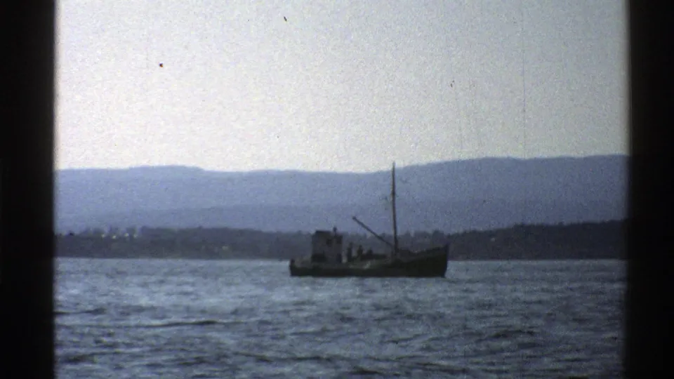 1976 Norway home-movie frame of a small boat on rippled water, backed by distant shoreline and blue-gray mountains.