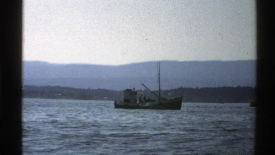 1976 Norway home-movie frame of a small boat on blue-gray water, backed by shoreline and hazy mountains.