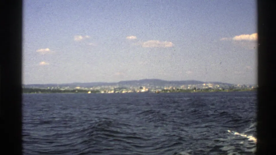 1976 Norway 8mm view across open water toward a distant coastal settlement and low hills under scattered clouds.