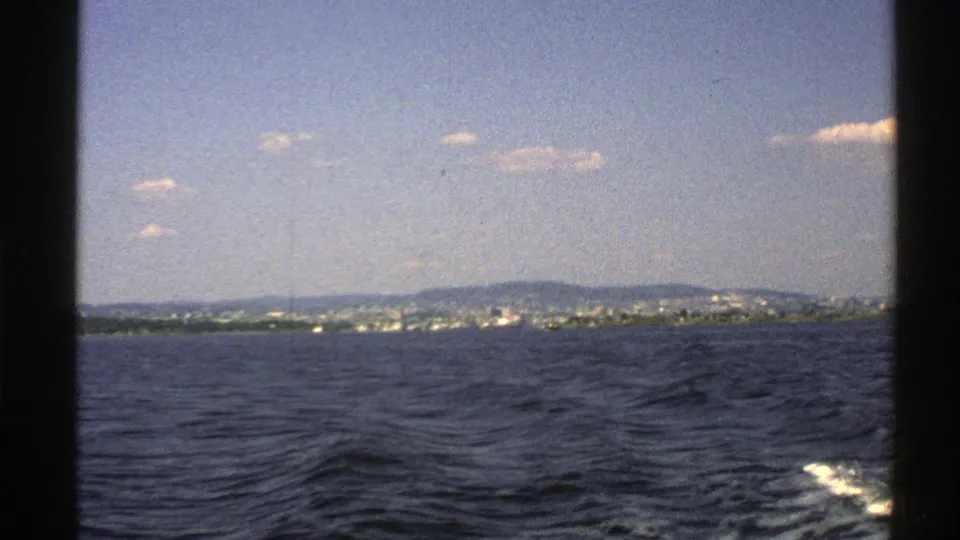 1976 Norway coastal home-movie frame with rippled water, distant buildings or shoreline, low hills, and a small wake at frame edge.