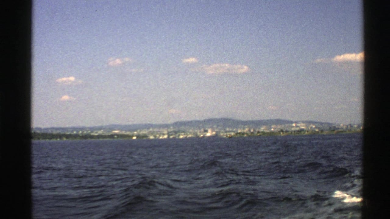 1976 Norway home-movie frame with dark boat framing, choppy water, white wake, shoreline buildings, hills, and small clouds.