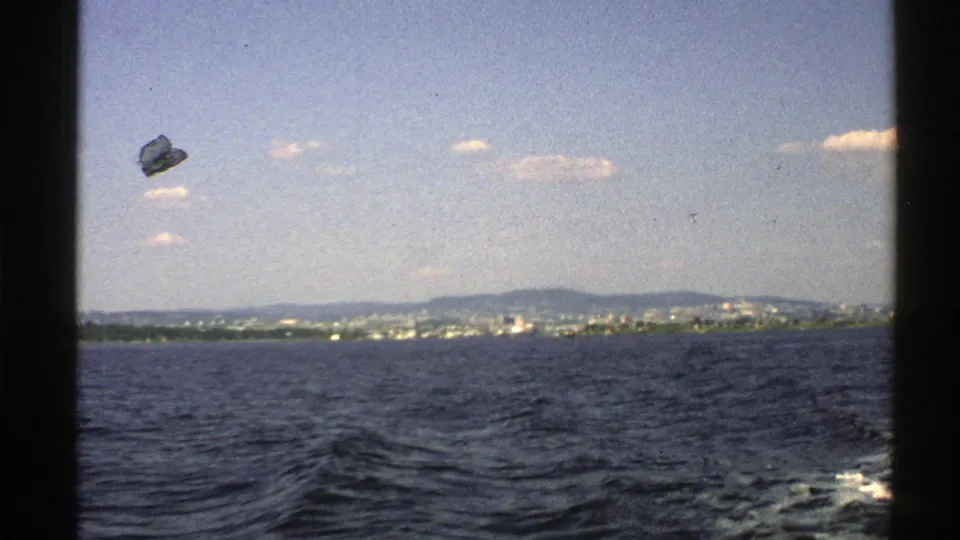 1976 Norway 8mm frame showing rippled water, a far shoreline with buildings, low hills, clouds, and visible film debris near the sky.