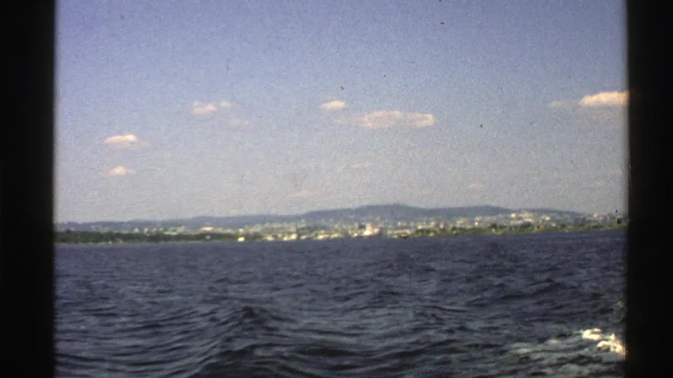 1976 Norway travel frame showing dark rippled water, a faint wake, distant waterfront buildings, low hills, and scattered clouds.
