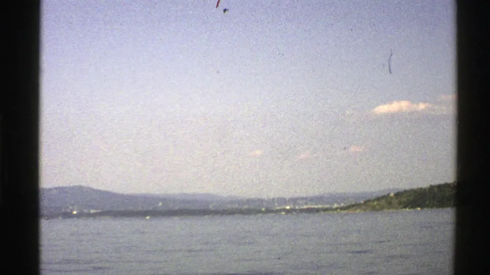 1976 Norwegian 8mm frame looking across calm water toward low hills and a distant shoreline, with film grain and gate edges visible.