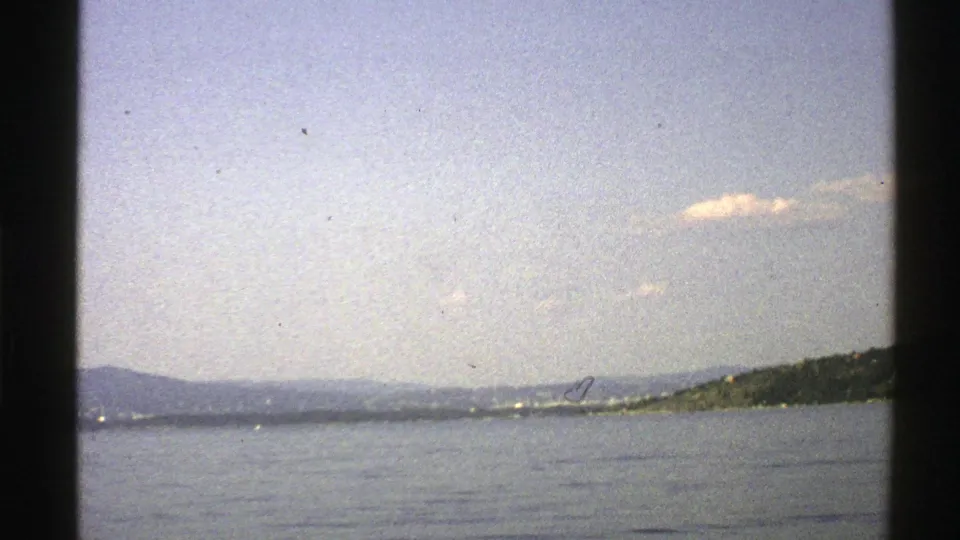 1976 Norwegian 8mm frame showing a calm water surface, low hills, distant shore buildings, and a wooded slope at right.
