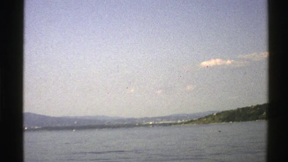 1976 Norway 8mm view across calm sea toward a distant tree-lined shore and low hills under a pale sky.