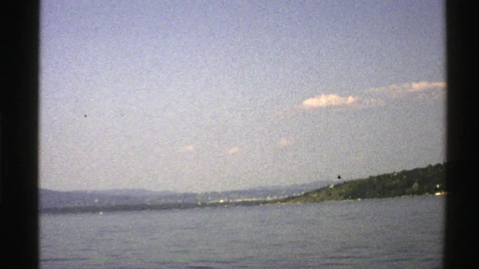 1976 Norway 8mm frame showing calm coastal water, a distant shore, low hills, and a small cloud above.