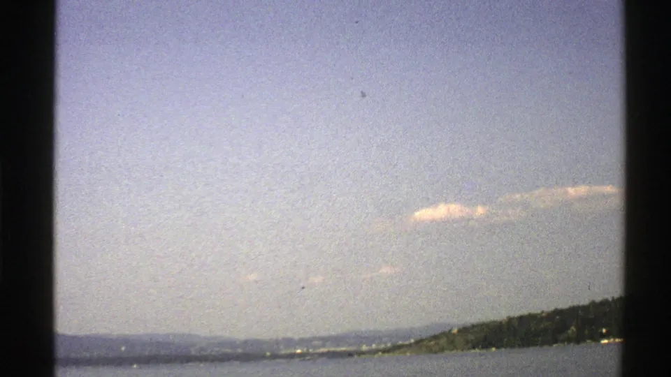 1976 Norway home-movie view of a broad sea surface, distant hills, and wooded coastline under pale blue sky.