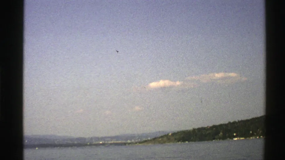 Faded 8mm view from Norway in 1976 showing open water, a distant wooded island, low hills, and small clouds overhead.