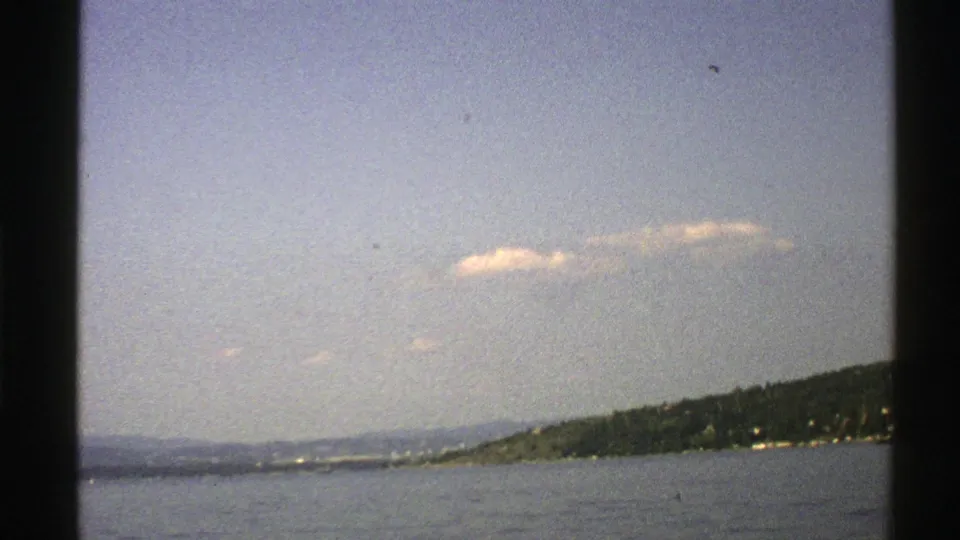 A 1976 8mm Norway coastal frame shows calm water and a wooded island shoreline at right beneath a pale clouded sky.