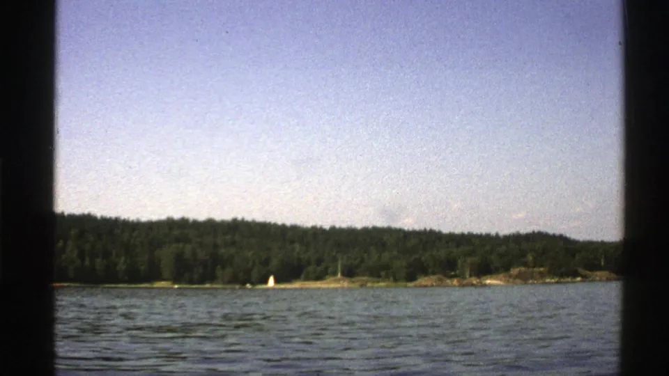 1976 Norway lake view with rippled blue water, wooded shore, and a small white sailboat under clear daylight.