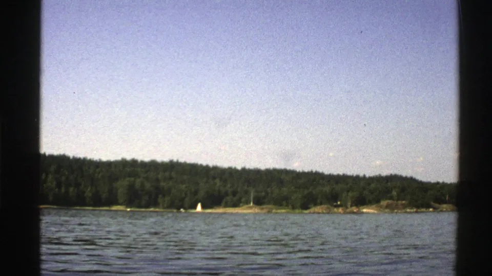 1976 Norway 8mm frame of rippling lake water, tree-covered shore, and a distant white sailboat in bright daylight.