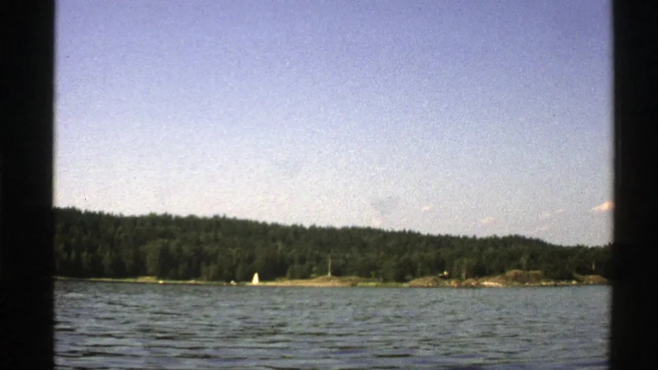 1976 Norway home-movie frame showing calm lake ripples, a forested shore, and a small white sailboat near the horizon.