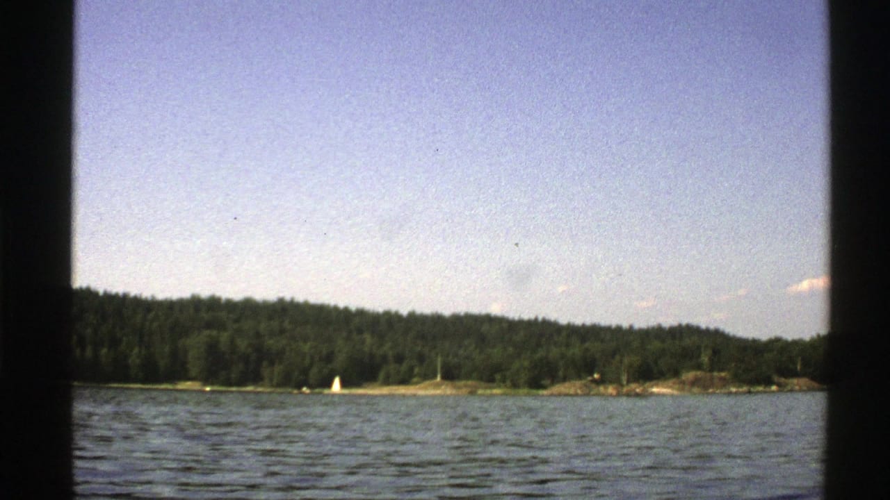 Boat-level view across rippled water toward a wooded shoreline and small white sailboat.