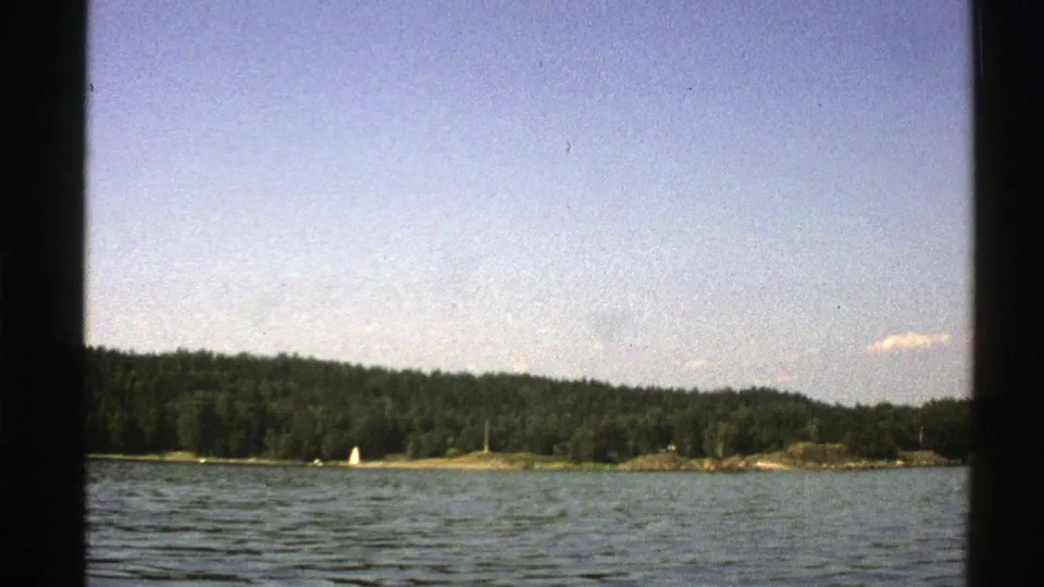 1976 Norway 8mm still showing open water in the foreground, a tree-lined shore, and a small white sailboat near the land.