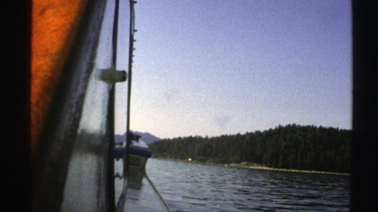 1976 Norway 8mm frame with the boat edge and pole in the foreground, open water, wooded shore, distant hills, and a small yellow shore light.