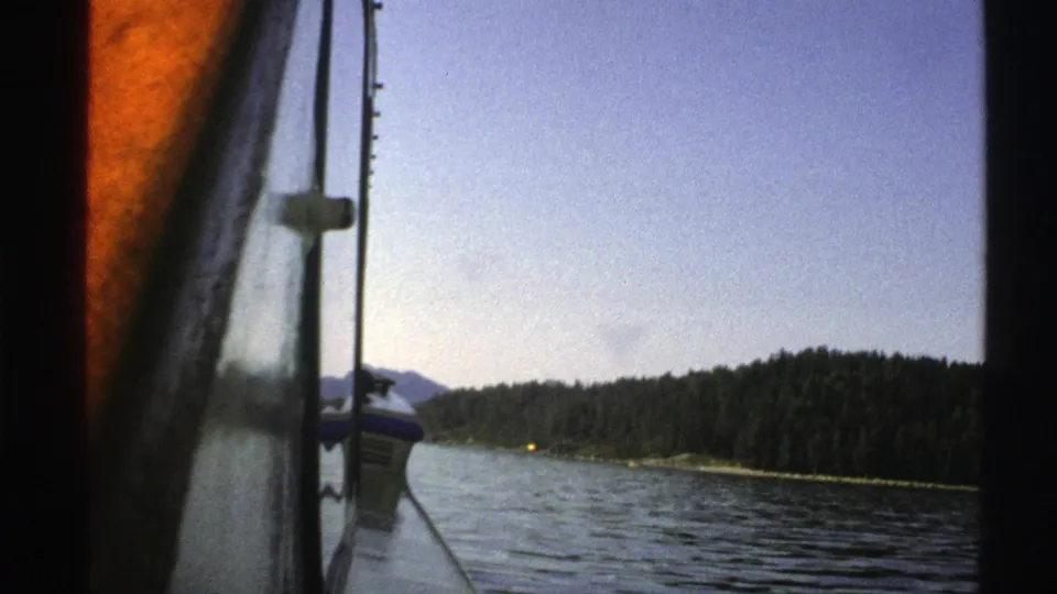 1976 Norway 8mm frame with the boat edge and pole in the foreground, open water, wooded shore, distant hills, and a small yellow shore light.