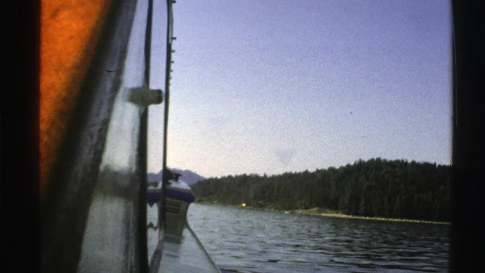 1976 Norway 8mm frame looking from the vessel across rippling water to a wooded shoreline, with distant hills and soft film grain visible.
