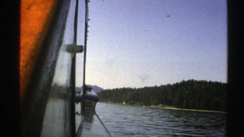 1976 Norway 8mm frame from inside a boat, with a vertical pole, water, wooded shoreline, distant hills, clear sky, and visible film specks.