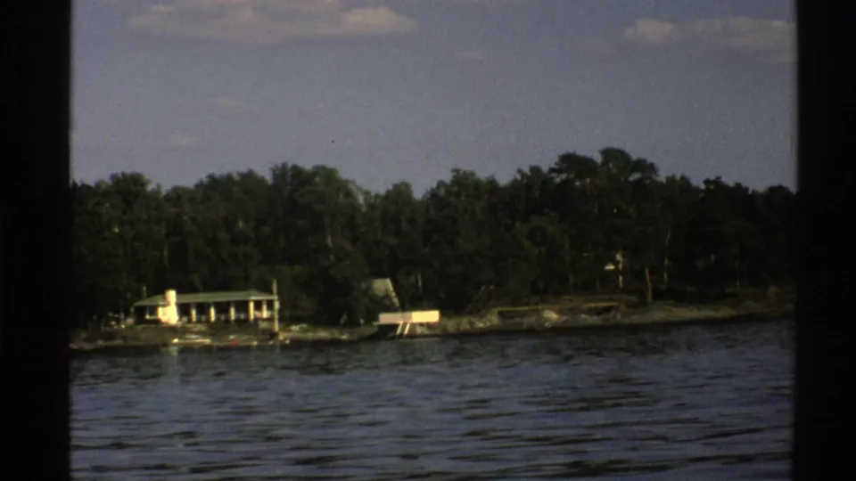 Norwegian lakeshore seen from a moving boat, showing rippling water, dense trees, a small building, and low dock-like structures.