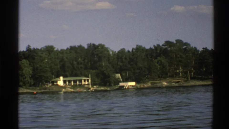 Late clip frame of a wooded Norwegian lakeshore from the boat, with a small light building, low waterfront structures, and rippled water.