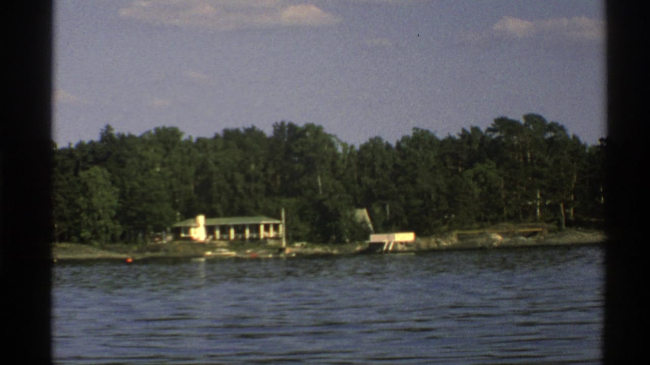 Light-colored lakeside house across calm water with forest behind it.